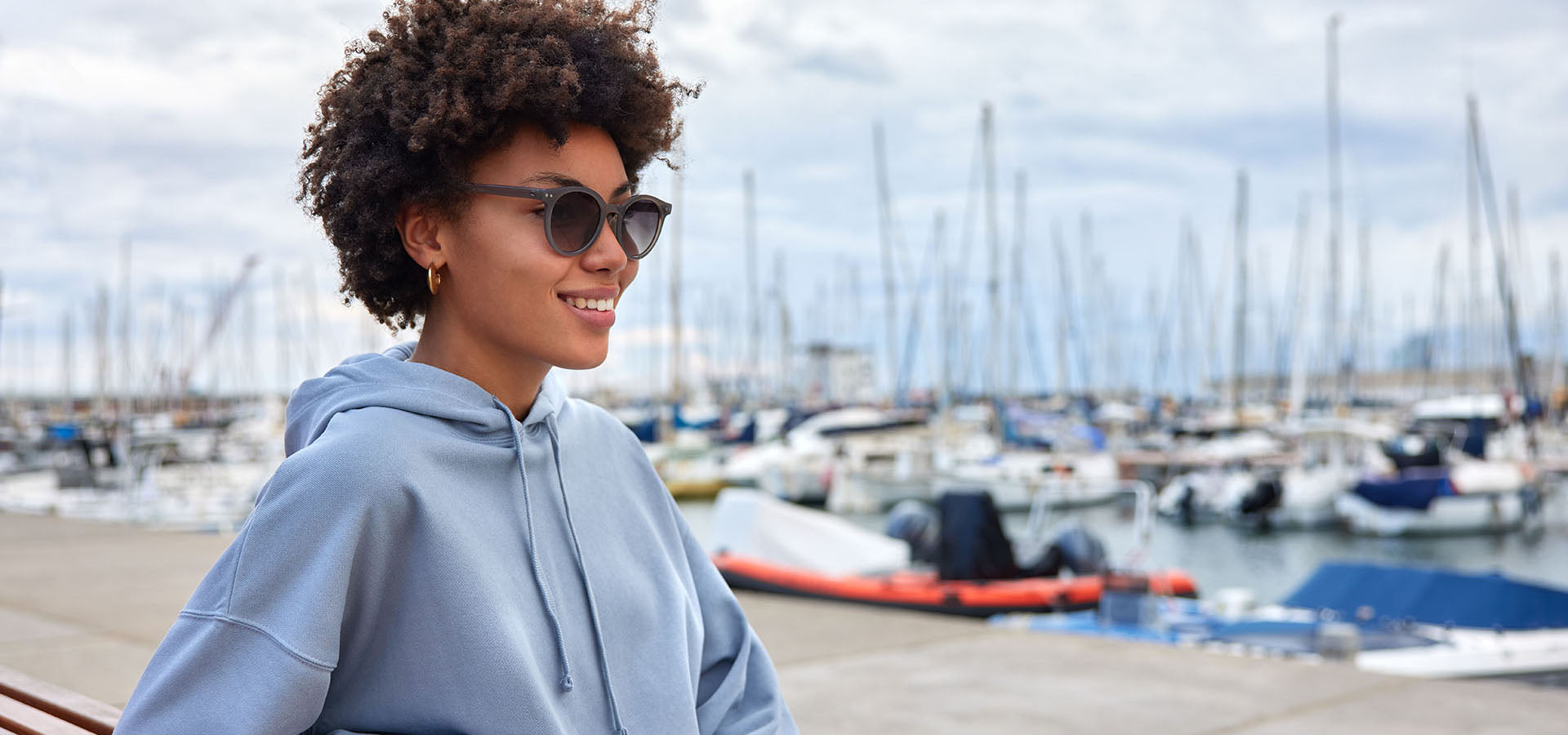Beautiful woman in a blue hoodie and sunglasses standing by a marina with boats in the background at accexo.com