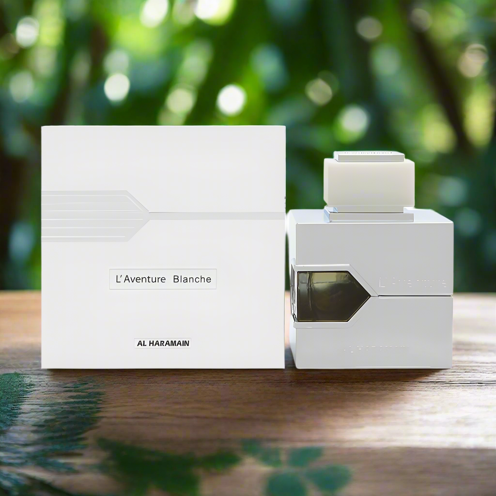 White perfume bottle and packaging with 'L'Aventure Blanche' branding on a wooden surface with green foliage in the background.